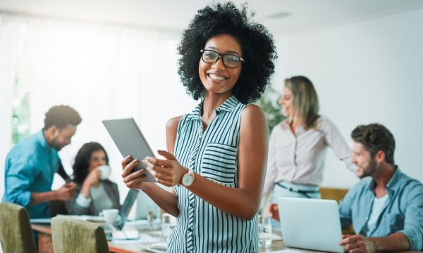 Black woman smiling and holding ipad stands in front of people working behind her